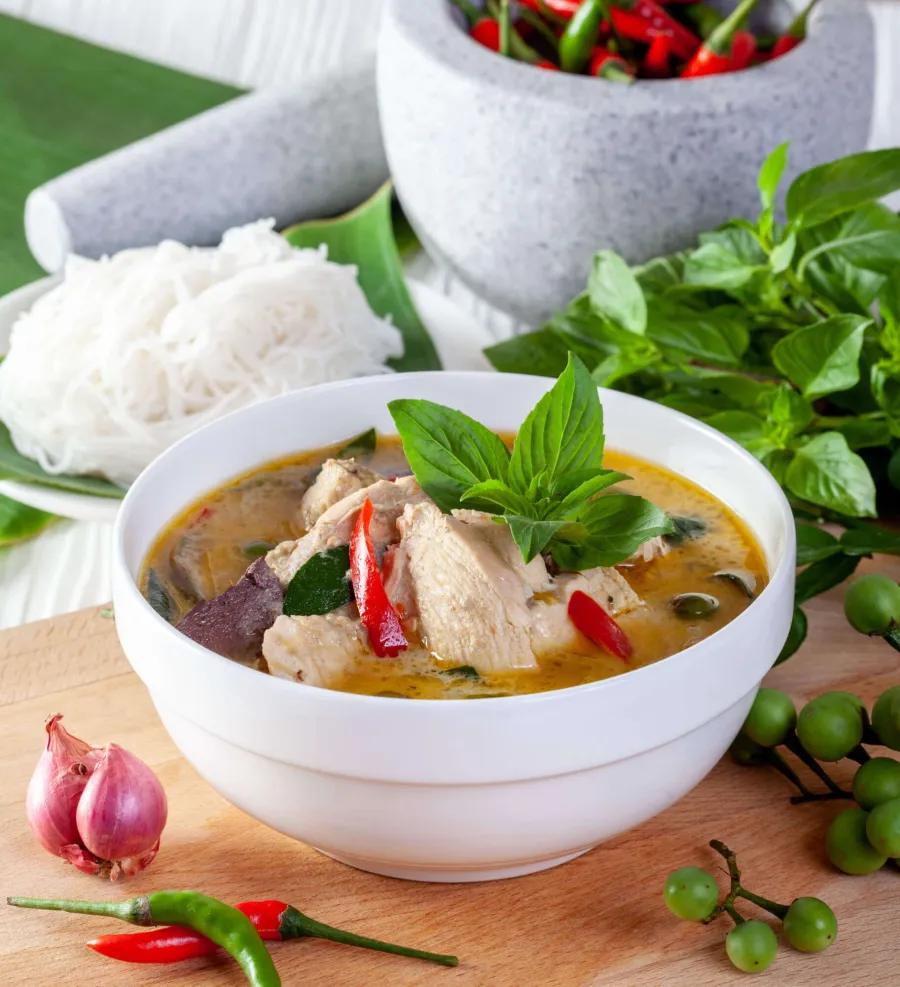 แกงเขียวหวานไก่ในชามขาววางคู่ขนมจีนและใบโหระพา